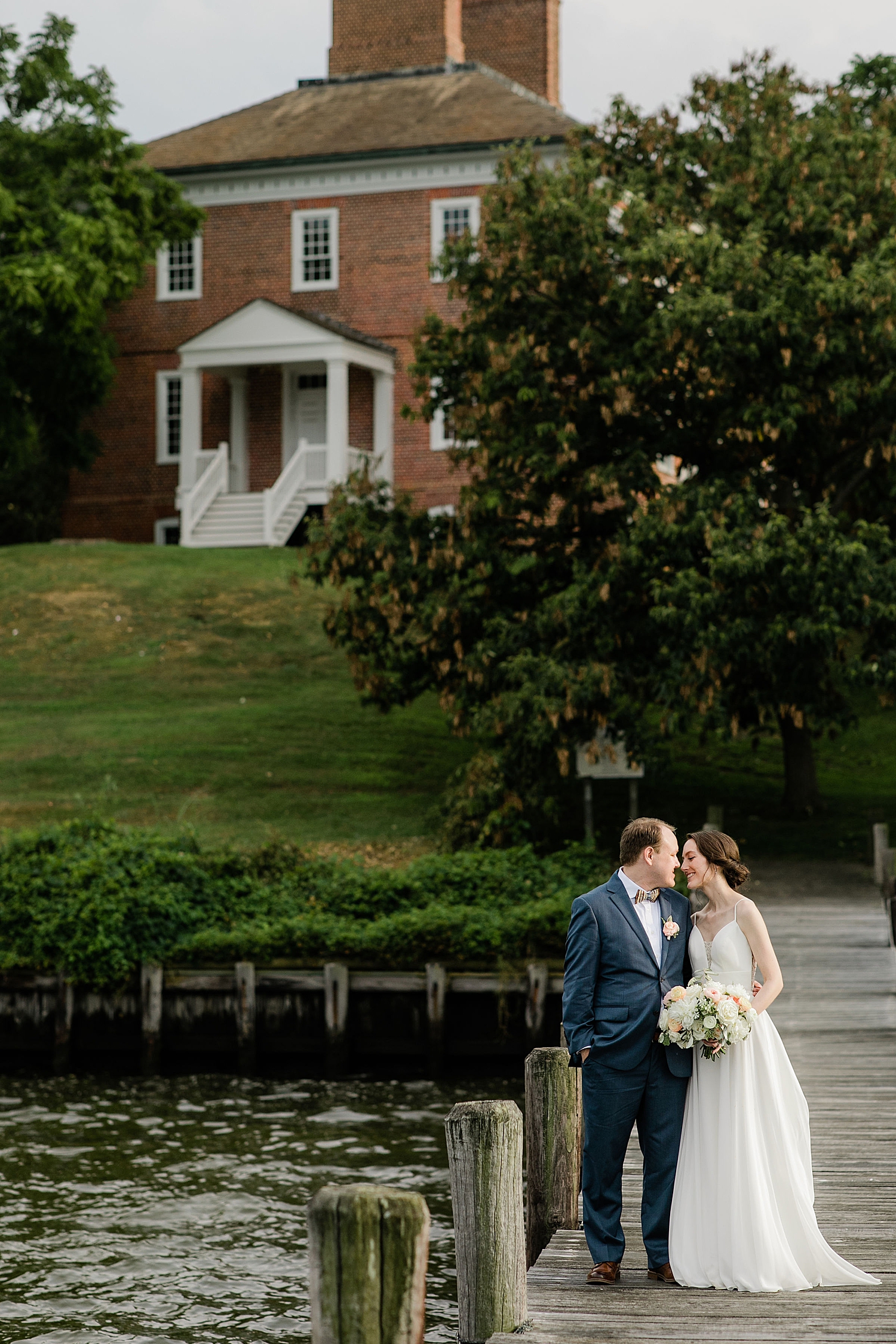 couple in formal wear embrace on dock with historic house behind them by Jen Harvey Photography