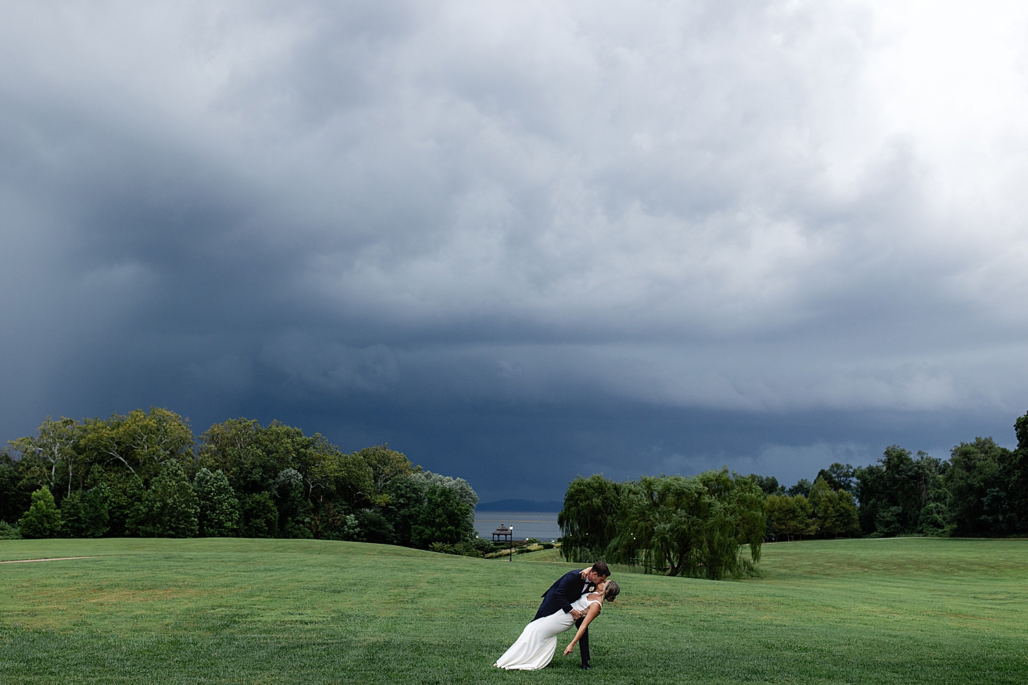 man dips his bride for a kiss in large open field in front of storm clouds at Swan Harbor