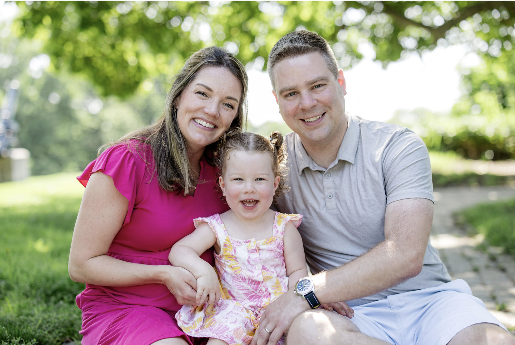 expecting parents and their toddler smiling together surrounded by soft greenery