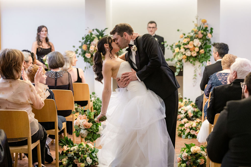 first kiss during wedding ceremony venue surrounded by peach and ivory floral arrangements