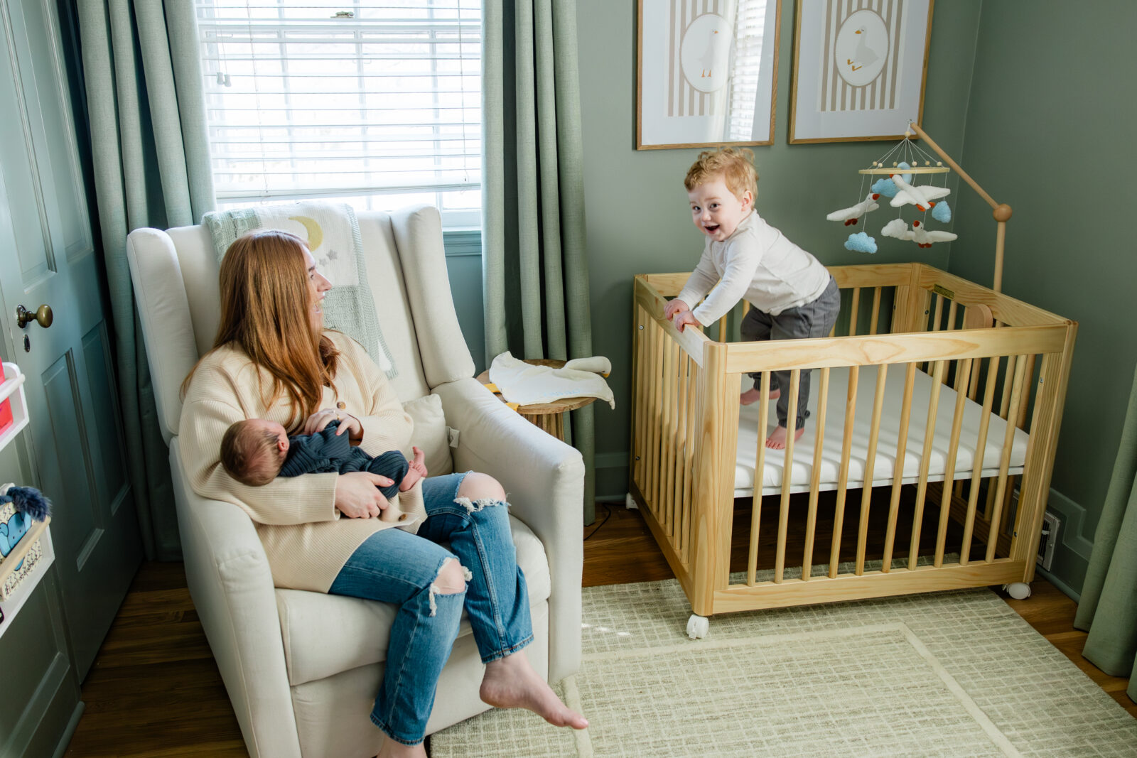 A mom holding a newborn son while her toddler son climbs in the crib.