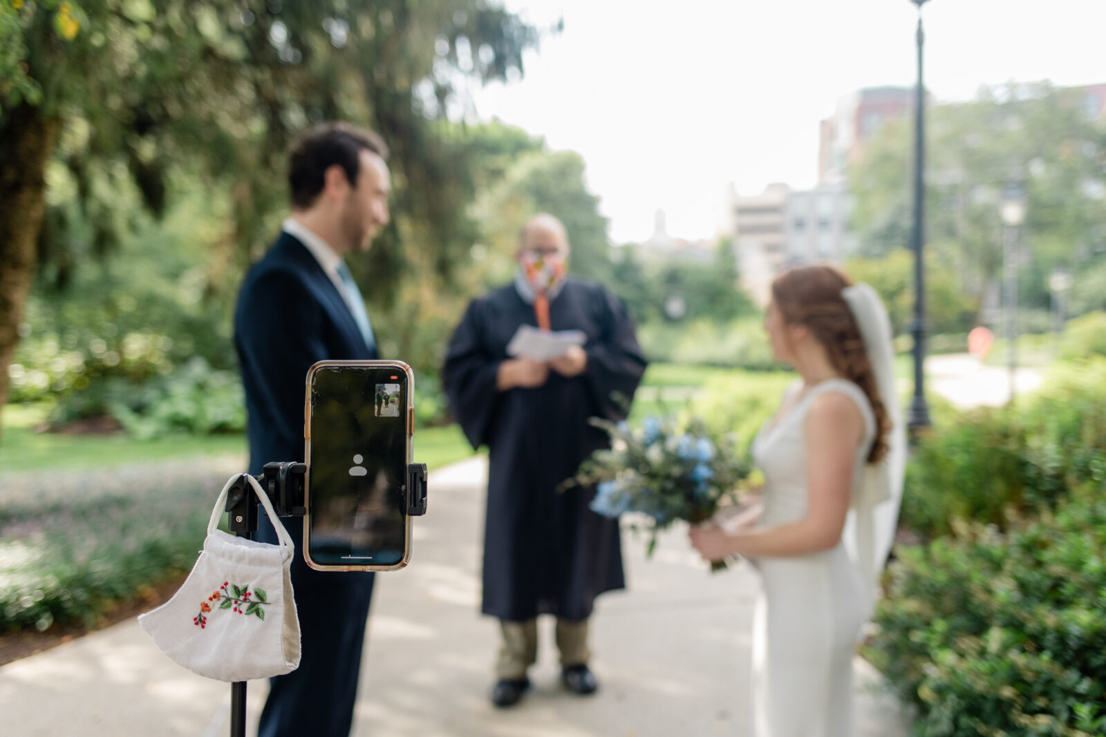 A bride and groom getting married being recorded on a phone with a face mask hanging off it.