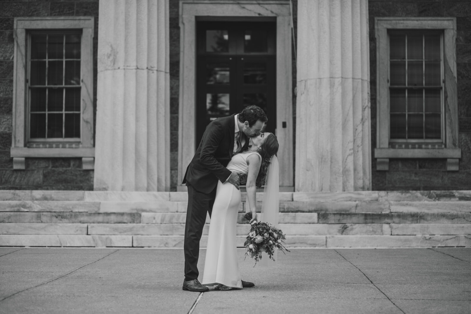 A black and white photo of a bride and groom kissing outside the courthouse.