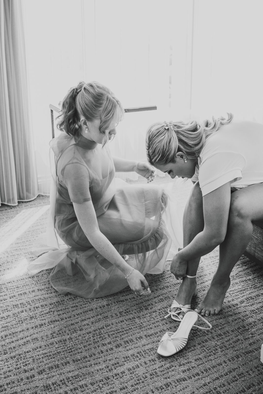 black and white photo showing bridesmaid helping bride put on her shoes